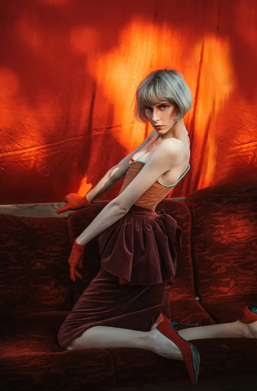 A model with a silver crop kneels on a red velvet sofa, glancing back over one shoulder. The gray laced corset from the original collection worn against a warm red-drenched backdrop — intimacy charged with editorial tension.