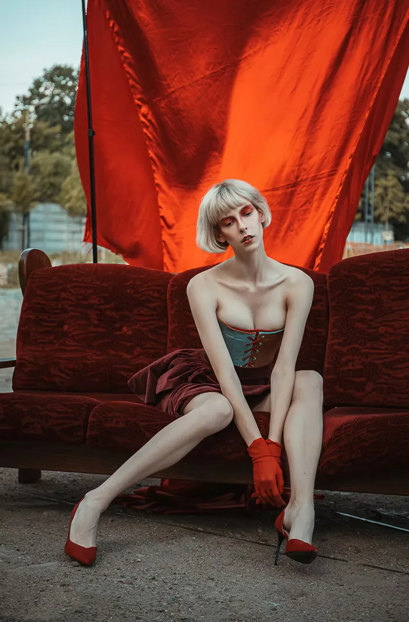 The gray corset and burgundy skirt, now worn outdoors on a red velvet sofa against draped red fabric. The original costume reframed as street-facing, public, fully in the world.