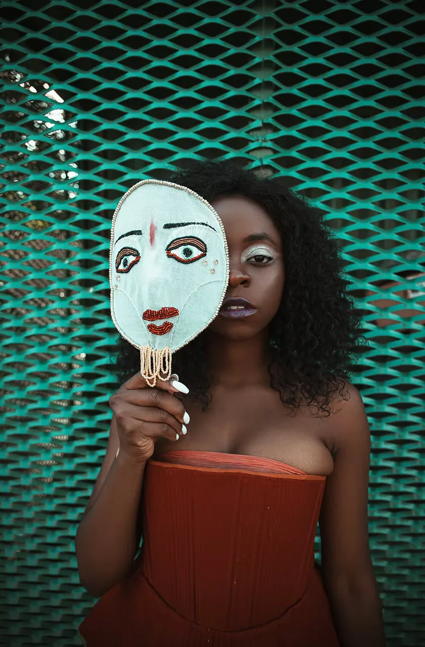 A Black model holds a hand-painted theatrical mask before their face against a teal chain-link fence, wearing a terracotta strapless bodice. The image foregrounds the project's central question: who wears the face of the Other, and who decides?
