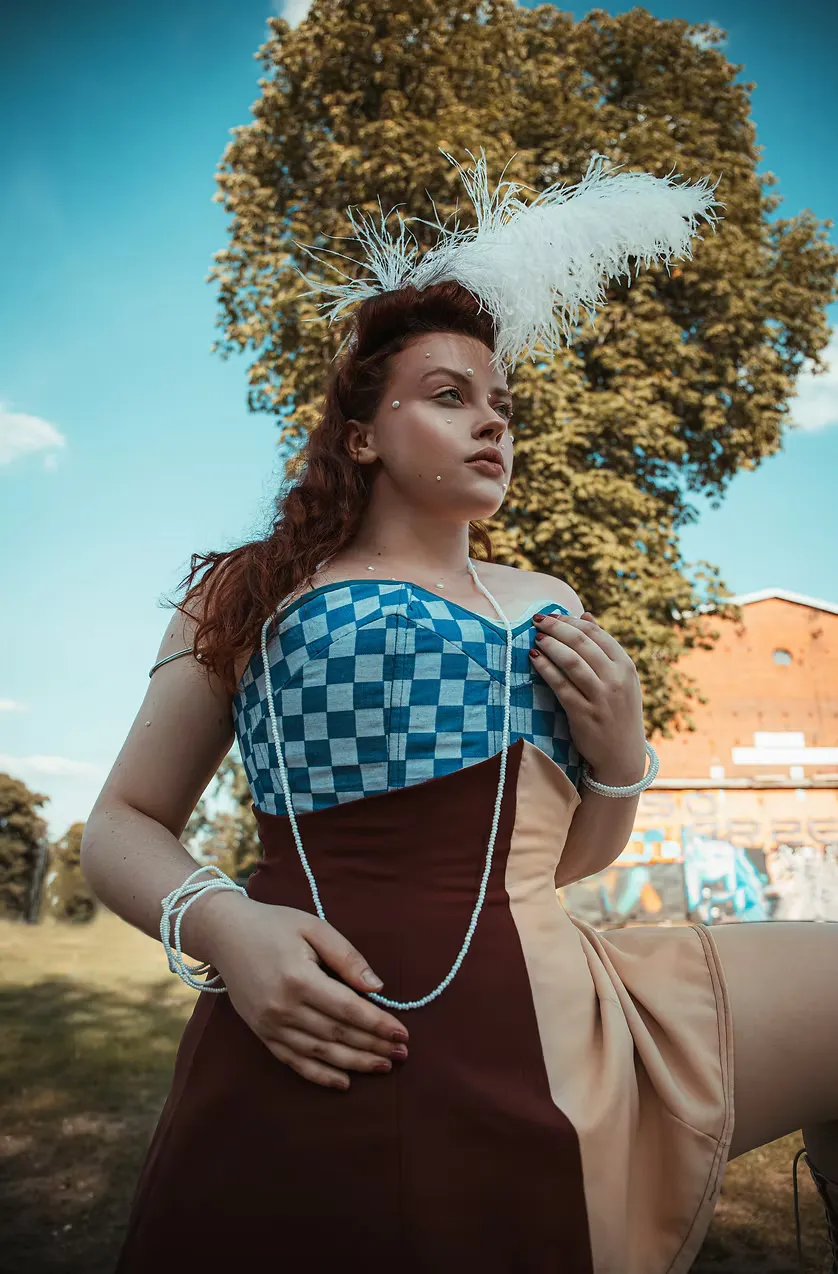A model with auburn hair stands outdoors in a blue checkerboard strapless bodice, brown skirt, and white feathered headpiece, pearl strands at the collarbone. The Courtesan character reimagined — ornate, grounded, and fully present in the world.