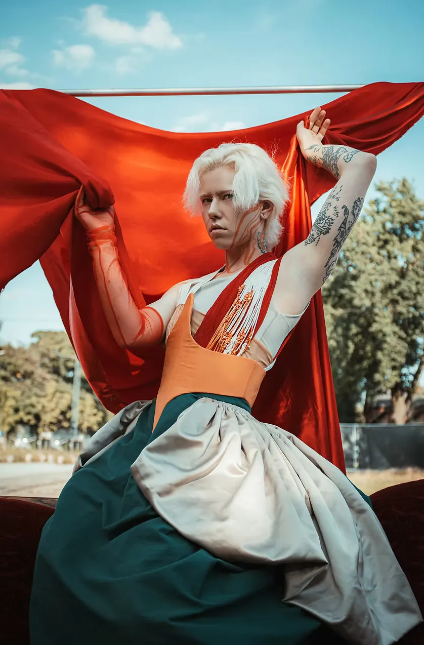 A model with short white hair and tattooed arms stretches red fabric wide against open sky, the teal-and-orange costume catching the daylight. A figure suspended between performance and freedom.