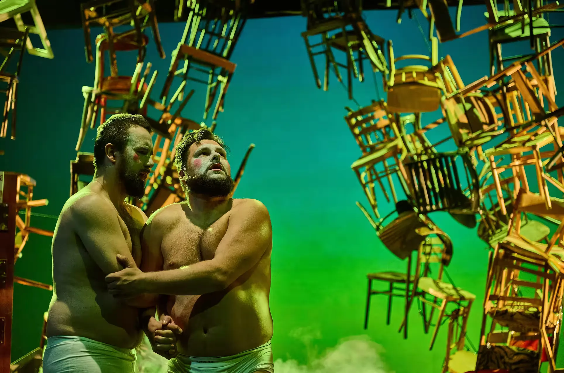 Two actors with white makeup and shirtless, holding each other