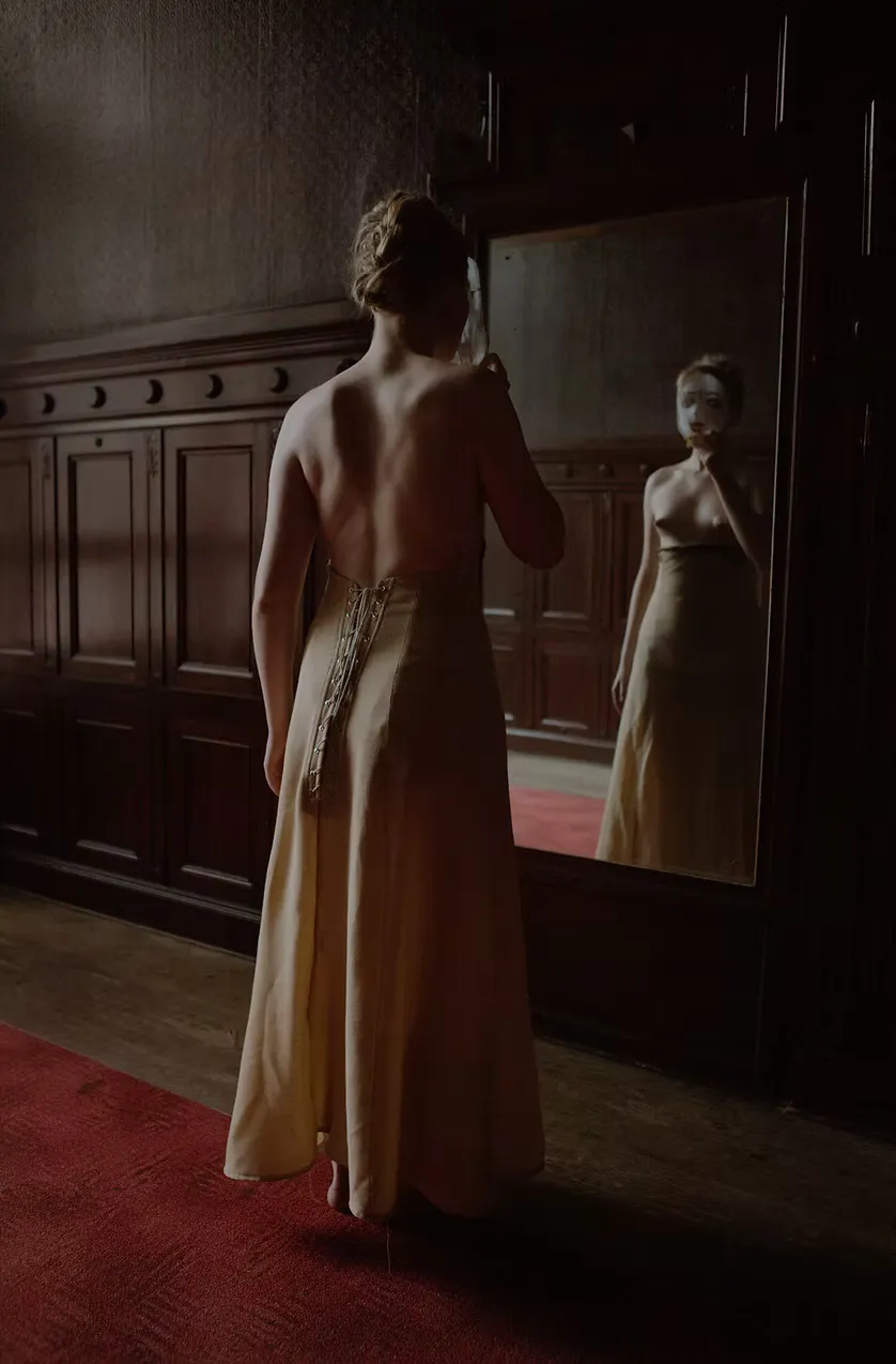 Full-length view of a figure in a backless pale gold gown, her reflection caught in an antique standing mirror within a dark interior. Red carpet anchors the composition. The self observed, the self constructed.
