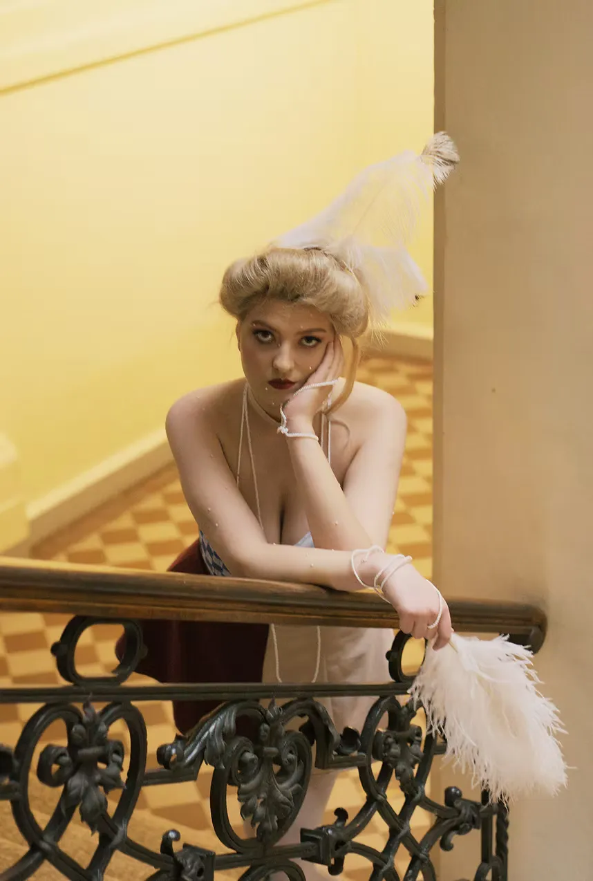 Leaning over a wrought-iron balcony rail against a warm ochre wall, a figure in a décolleté cream dress holds an ostrich feather fan. A small feathered ornament frames the face. The courtesans of fin-de-siècle Europe — at once ornamental and sovereign.