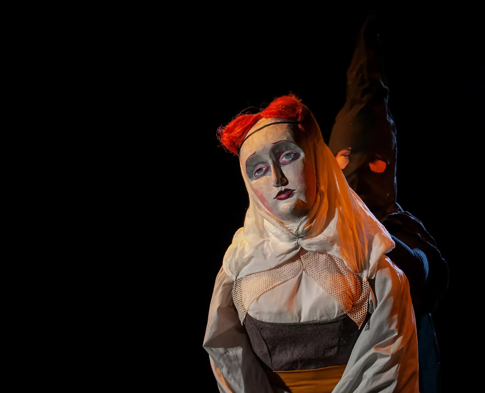 On a dark stage, a white-faced puppet figure with heavy theatrical makeup, red hair, and a ruffled white collar stands before a looming hooded figure in black. Commedia dell'arte atmosphere, shadow and theatre intertwined.