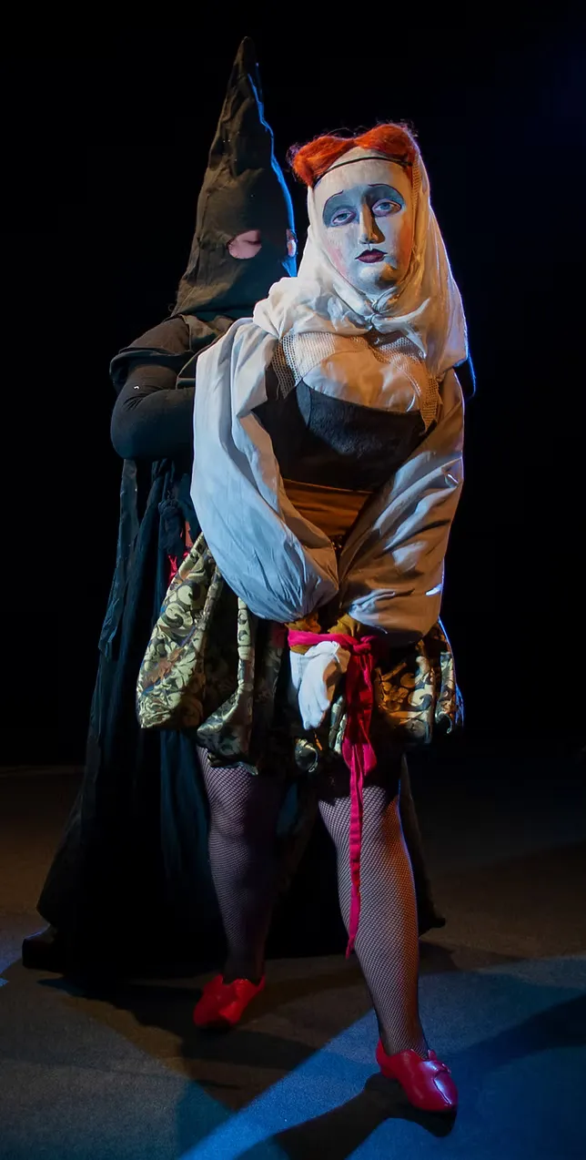 Full-stage view of a white-faced puppet figure in a white blouse and amber bodice, accompanied by a hooded puppeteer in black. Red shoes catch the stage light. Theatrical and quietly uncanny.