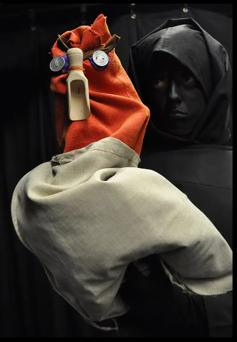 The large golden-masked puppet in black robes seen from a closer angle on the metal scaffolding. The mask's dramatic painted expression fills the frame.