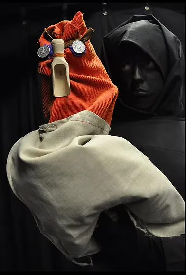A large puppet in black robes and a round golden mask with dramatic painted eyes grips the bars of a metal scaffolding structure in an exhibition space. Commanding and theatrical in scale.
